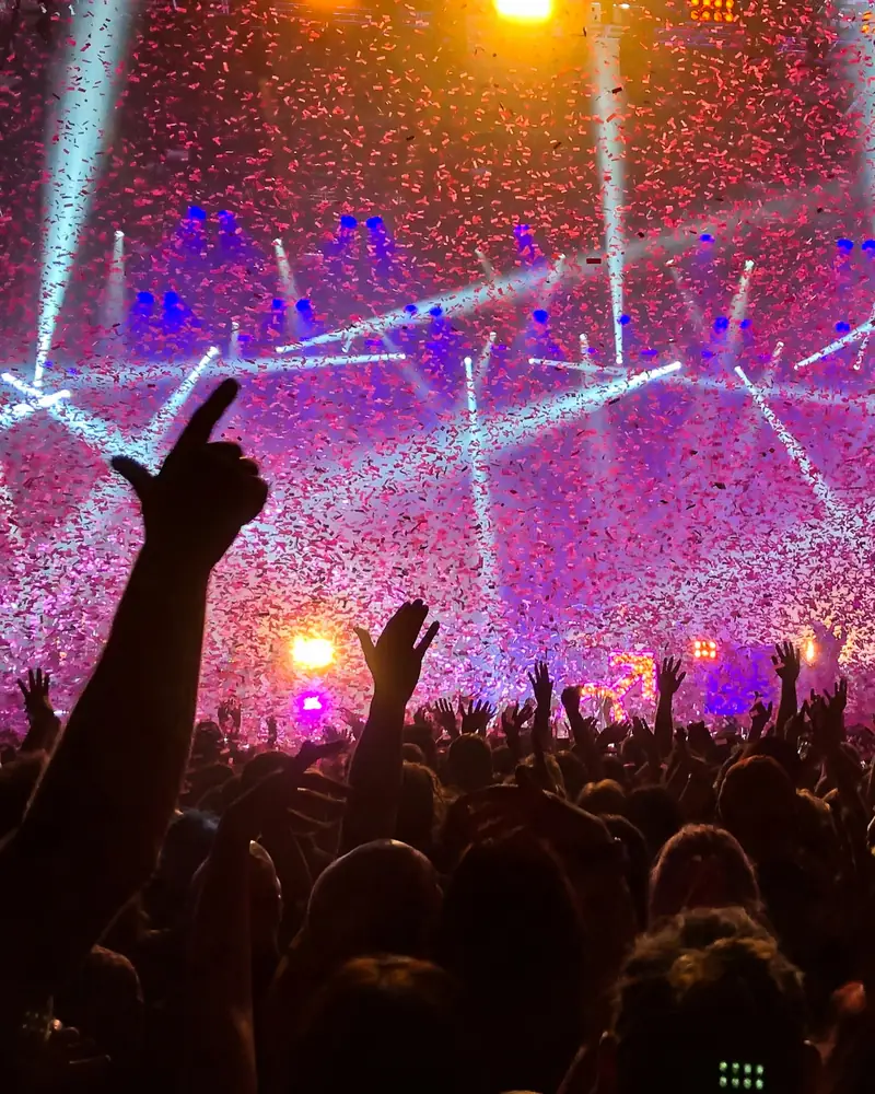 A crowd with hands in the air as confetti and lasers fill the room.